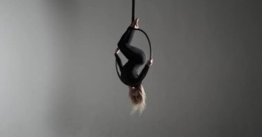 Kendine güvenen beyaz bir dansçı havadaki çemberde cazibesine kapılır ve akrobatik yetenekleriyle yer çekimine meydan okur. Karşıt ışıklarla aydınlatılmış, karanlık bir arkaplanda.