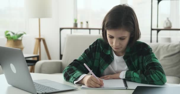 少年は居間のテーブルに座っている 彼はノートパソコンを使ったオンラインレッスンに積極的に参加している 彼の顔は興味を反映し 彼の手はノートに書いて忙しい — ストック動画