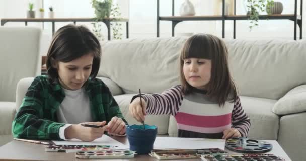 テーブルの近くの床には白人の兄弟姉妹が座っていて 楽しいアートセッションをしています 鉛筆と水彩画を手にして 彼らは創造的なプロセスに没頭します — ストック動画