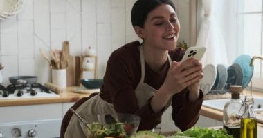 Mutfakta beyaz bir kadın var. Telefona dalmış, kaseden lezzetli salatanın tadını çıkarıyor. Rahatlamış tavır ve çoklu görev gösterimi mutfak zevkleri ve dijital bağlantı ile modern yaşam tarzı.