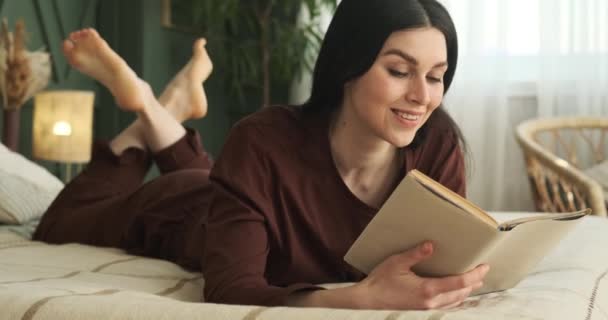 居心地の良いパジャマ姿の白人女性がベッドの上に横たわっていた 魅力的な本を手に 彼女はそのページに飛び込みます 彼女は広く笑顔として顔は純粋な喜びを放射します — ストック動画