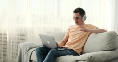 Kafkasyalı bir adam kanepeye oturmuş, kulaklık takmış ve bilgisayarındaki bir video görüşmesine dalmış. Odaklanmış bir ifadeyle, sanal dünyada başkalarıyla bağlantı kurmak için modern teknolojiyi kullanıyor..