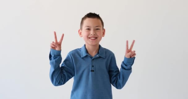 亚洲学童在白色的背景上表现出标志性的胜利姿态 这个小男孩面带微笑 伸出手指 构成胜利的标志 散发着积极和热情 — 图库视频影像