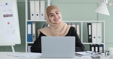 Kendine güvenen genç bir Arap iş kadını, tesettür takmış, kameraya bakıyor, ofis masasında otururken pozitiflik saçıyor. Parlak gülüşü iyimser bakışını ve bulaşıcı coşkusunu yansıtıyor..