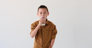 Asyalı okul çağındaki çocuk beyaz bir arka fonda duruyor ve bir bardak sudan ferahlatıcı bir yudum alıyor. Soğukluğu ve susuzluğunu giderirken yüzü mutluluk saçıyor..