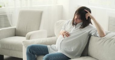 Sıcak bir oturma odasında, hamile bir kadın kanepede oturur, düşüncelere dalmış bir şekilde. Düşünceli bir ifadeyle, karnını nazikçe okşar, annelik yolculuğunu düşünür..