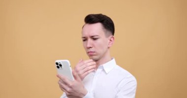 Beyazların bej arka planında yaşayan beyaz bir adam, dikkati dijital alemde. Konsantrasyon ifadesinde, bir telefon kullanıyor, arayüzü sorunsuz bir şekilde kaydırıyor ve kaydırıyor..