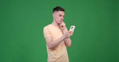 Yeşil arka planda ciddi bir beyaz adam, telefonunu canlı yeşil bir arkaplanda kullanıyor. Odaklanmış bir ifadeyle dikkatini ekrana veriyor..