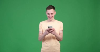 Esinlenmiş beyaz bir adam yeşil arka planda telefonunu kullanıyor. Yüzü coşkuyla parlıyor ve gözleri heyecandan parlıyor. Telefonu kendine güvenerek tutuyor..