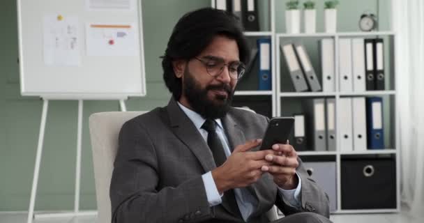 眼鏡をかけたインド人ビジネスマンとネクタイをしたスーツがオフィスの快適なアームチェアに座り 笑顔で電話をかけている 生産的なコミュニケーションに従事する起業家 — ストック動画