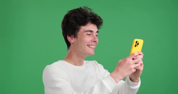 在一个充满活力的绿色背景下 他热情地在手机上打字 微笑着白人男子 他以愉快的表情和生动的动作流露出一种积极和愉快的感觉 — 图库视频影像