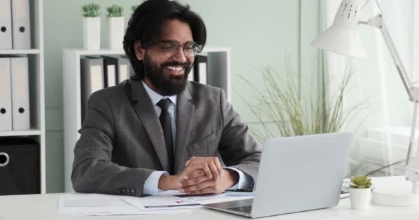 インドのビジネスマンがオフィスの机に座っていて 友人とビデオ通話をしている 彼は彼らとチャットしながら 彼らが言う何かに笑顔で幸せそうです — ストック動画