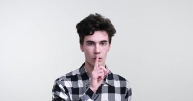 Beyaz bir arka plana karşı duran beyaz bir adam. Ciddi bir havayla, sessiz kalma ve otorite mesajını iletmek için parmağını dudaklarına kaldırır..
