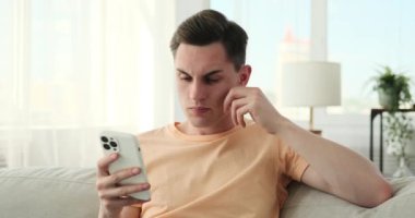 Bir adam koltukta oturuyor, elinde cep telefonu var ve şaşkın bir ifade takınıyor. Kaşları çatılmış, ve dikkatle telefon ekranına bakıyor, bir şeyi anlamaya çalışıyor..