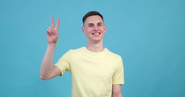 一个有着蓝色背景的快乐的高加索人举起他的手 形成了一个标志性的V形手势 通常被称为胜利的标志 脸上挂着大大的笑容 — 图库视频影像