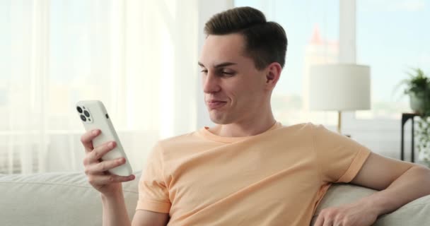 年轻的白人男子坐在沙发上 在家里拿着手机和信用卡享受网上购物 他面带微笑 对方便的购物方式表示高兴和满意 — 图库视频影像