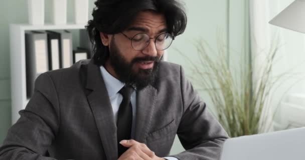 一位身穿西装 打着领带的印度商人坐在办公室的笔记本电脑前 一边微笑着 一边举手 一边与同事们分享他在视频通话中取得的成功 — 图库视频影像