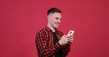 Canlı kırmızı arka planda neşeli beyaz bir adam telefonunda neşeli bir mesajlaşma sohbeti ile meşgul. Yüzü parlak ve bulaşıcı bir gülümsemeyle süslenmiş..