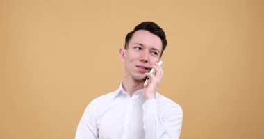 Sakin bir bej arka planda duran beyaz bir adam. Telefonunu kullanıyor ve yüzünde neşe dolu bir gülümsemeyle sohbet ediyor. Yüzündeki ifade pozitif bir havayı ortaya çıkarıyor..
