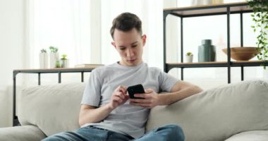 Şık dekore edilmiş bir oturma odasındaki koltukta oturan düşünceli beyaz adam. Düşünceli bir tavırla, telefonuna dalmış, mesajlaşırken ya da içeriği karıştırırken..