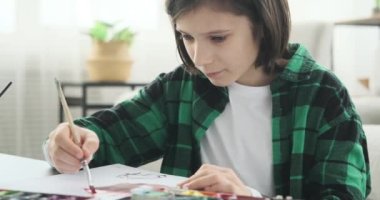 Yetenekli beyaz çocuk evinde bir masanın yanında rahat bir şekilde oturuyordu. Yaratıcı görüntülerini renkli suluboya tabloları kullanarak hayata geçiriyordu..