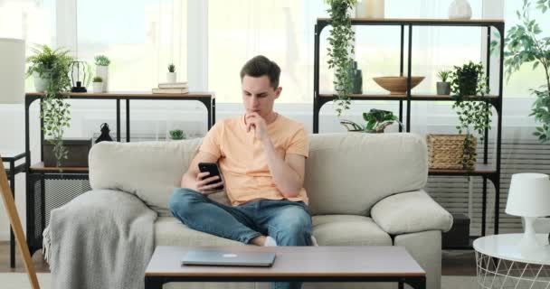 一个心事重重的白人男人坐在舒适的沙发上 沉浸在智能手机里 带着沉思的表情 他在数字世界中航行 陷入沉思 — 图库视频影像
