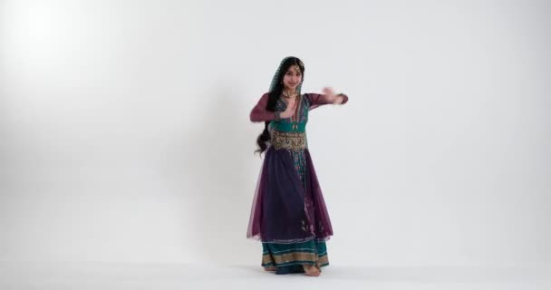 一位年轻的印度妇女在传统音乐的伴奏下 在白色的背景上翩翩起舞 身穿蓝色 紫色和绿色的色彩艳丽的莎丽服 装饰着传统的印度珠宝 — 图库视频影像