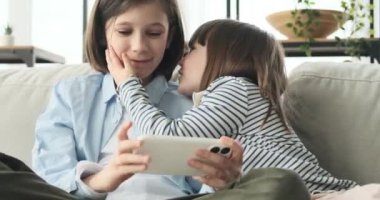 İki kardeş, bir erkek kardeş ve bir kız kardeş, rahat bir koltukta oturuyorlar, teknoloji dünyasına dalmışlar, yüzlerinde geniş bir gülümseme olan bir telefon kullanıyorlar..