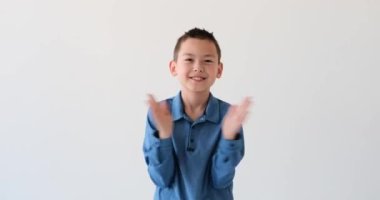 Yakışıklı Asyalı çocuk el değmemiş beyaz bir arka planda alkışlarken mutluluk saçıyor. Yüzünü aydınlatan ışıl ışıl bir gülümseme ile ellerini ritim ve coşkulu bir şekilde çırpıyor..