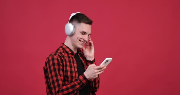 赤い背景には 幸せな白人男性がヘッドフォンをして携帯電話を持って描かれています 彼はヘッドフォンを通して音楽を聴くと喜びを放射する — ストック動画
