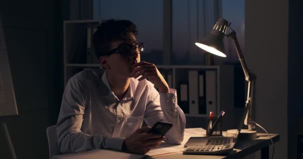 メランコリック コーカサス事務所の従業員が携帯電話を使っている 薄暗い照明は遅い時間を強調する一方 彼のダウンキャスト表現は悲しみや熟考の感覚を反映しています — ストック動画