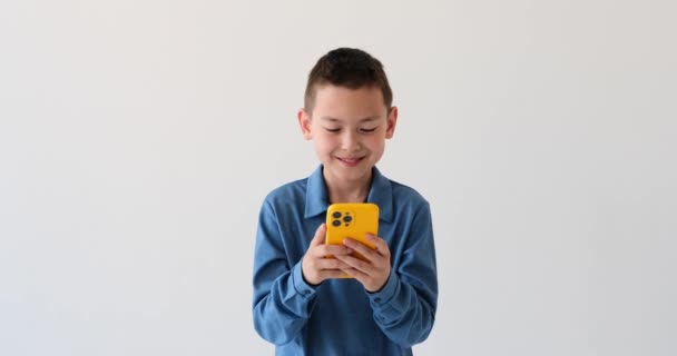 白い背景に学校の年齢の楽しいアジアの少年 彼の顔に伝染性の笑顔で 彼はデバイスに魅了され 彼は楽にスワイプし タップし さまざまなアプリやコンテンツを探検しました — ストック動画