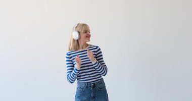 Sarışın bir kadın temiz beyaz bir arka planda, kulaklık takmış ve coşkuyla dans ediyor. Hareketleri ve neşeli ifadesi enerji ve mutluluk saçıyor..
