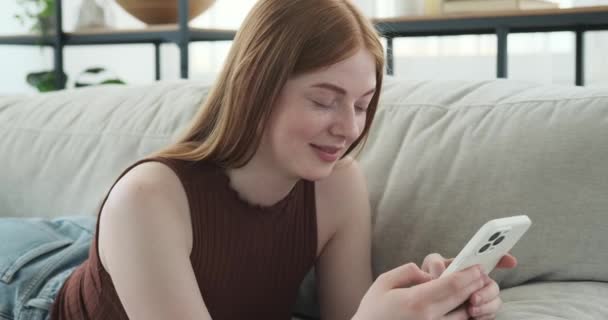 一个红头发的白人女孩躺在沙发上 用手机发短信 她脸上带着平静的表情 在周围环境中显得安详而舒适 — 图库视频影像