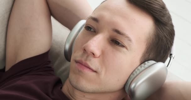 在这个无忧无虑的时刻 一个年轻人躺在沙发上休息 用耳机听音乐 他的脸上露出一副无忧无虑 心满意足的表情 — 图库视频影像