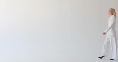 Sarışın bir kadın, beyaz bir zeminde profesyonel bir takım elbise giymiş, kasıtlı bir adımla geçerken görülüyor. Kendine güveni ve sakin tavırları hareket ederken amacını gösteriyor..