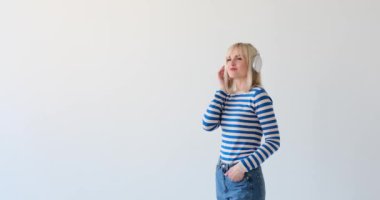 Beyaz kadın temiz beyaz bir arka planda, kulaklık takıyor. Müzik dinliyor, podcast yapıyor ya da sesle ilgili herhangi bir aktiviteye katılıyor olabilir..