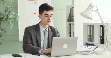 Ofiste bilgisayarını kullanan yakışıklı bir ofis çalışanı. Profesyonel görünüşü ve kendine güveni ile, dizüstü bilgisayarı üzerinde özenle çalışır, görevleri kolaylıkla yerine getirir..