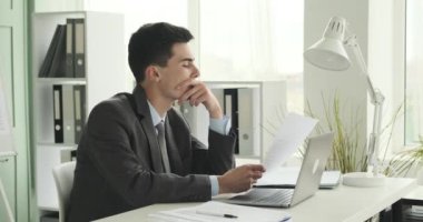 Genç ve odaklanmış beyaz adam ofisinde özenle çalışırken görülüyor. Masasında oturuyor, görevlerine dalmış, bir dizüstü bilgisayar açık, çeşitli belgeler ve belgeler önüne serilmiş..