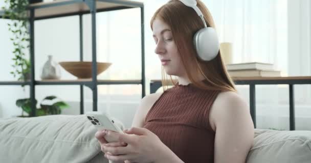 人们看到少女戴着耳机坐在沙发上 专心致志地听着手机 她看起来很专注 沉浸在她的数码世界里 可能是在听音乐 — 图库视频影像