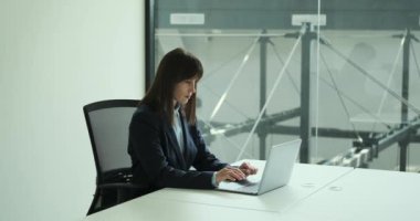 Çağdaş bir ofis ortamında, profesyonel beyaz bir iş kadını masasında oturmuş, özenle dizüstü bilgisayarı üzerinde çalışıyor. Odaklanmış bir ifadeyle, görevleri etkili bir şekilde yönetir..