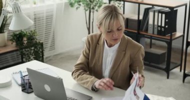 Ciddi ve odaklanmış iş kadını, belgelerdeki bilgileri bilgisayarındaki bilgilerle titizlikle karşılaştırırken resmediliyor. Kararlı bir ifadeyle, titizlikle verileri çapraz referanslar..