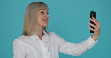 Güzel bir kadın, sakin bir mavi arka planda telefonla görüntülü konuşma yapıyor. Işıl ışıl gülüşü ve nişanlı ifadesi telefonun diğer ucundaki kişiyle olan eğlencesini ve bağlantısını yansıtıyor..