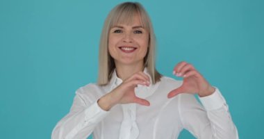 Sevgi dolu, beyaz bir kadın, sakin mavi bir arka planda elleriyle kalp şekli oluşturuyor. Sevecen ifadesi ve kalp hareketi aşk ve romantizm duygularını yansıtıyor..