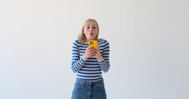 Sarışın kadın temiz beyaz bir arka planda elinde bir telefonla gerçekten şaşırmış görünüyor. Şaşkın bakışları ve kaşlarını kaldırması az önce gördüğü bir şeye olan şaşkınlığını gösteriyor..