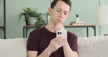 Derin düşüncelere dalmış, beyaz bir adam koltukta oturuyor, derin düşüncelere dalmış, telefonunu kullanıyor. Odaklanmış ifadesi anlamlı bir içeriğe daldığını gösteriyor..