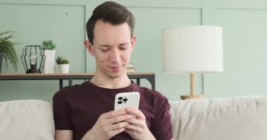 Bir adam kanepede otururken telefonunda gülerken ve mesajlaşırken görülüyor. Yüzü neşeli bir gülümsemeyle aydınlanıyor, neşeli havasını yansıtıyor..