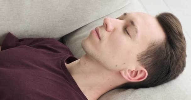 若い白人男性はリビングルームのソファーで眠っているように見えます 彼の顔は穏やかな表情を示し 耐え難いスラムを示しています — ストック動画