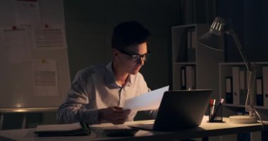 Stresli ofis çalışanı bir yığın belgeyle boğuşurken görülüyor. Masa lambasının yumuşak parıltısı gecenin geç saatlerine ve karşılaştığı zorluklara vurgu yapıyor..
