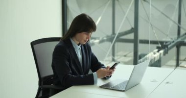 Ofiste kendine güvenen, profesyonel takım elbiseli bir kadın kendine güvenen bir şekilde telefonundan mesaj atıyor. Düzenli bir tavır ve odaklanmış bir ifadeyle, iletişimini etkili bir şekilde idare ediyor..